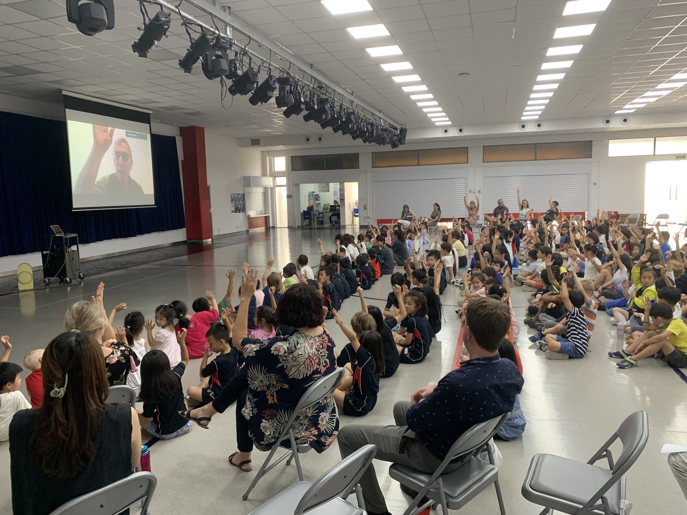 crowd of children raising hands at man on screen