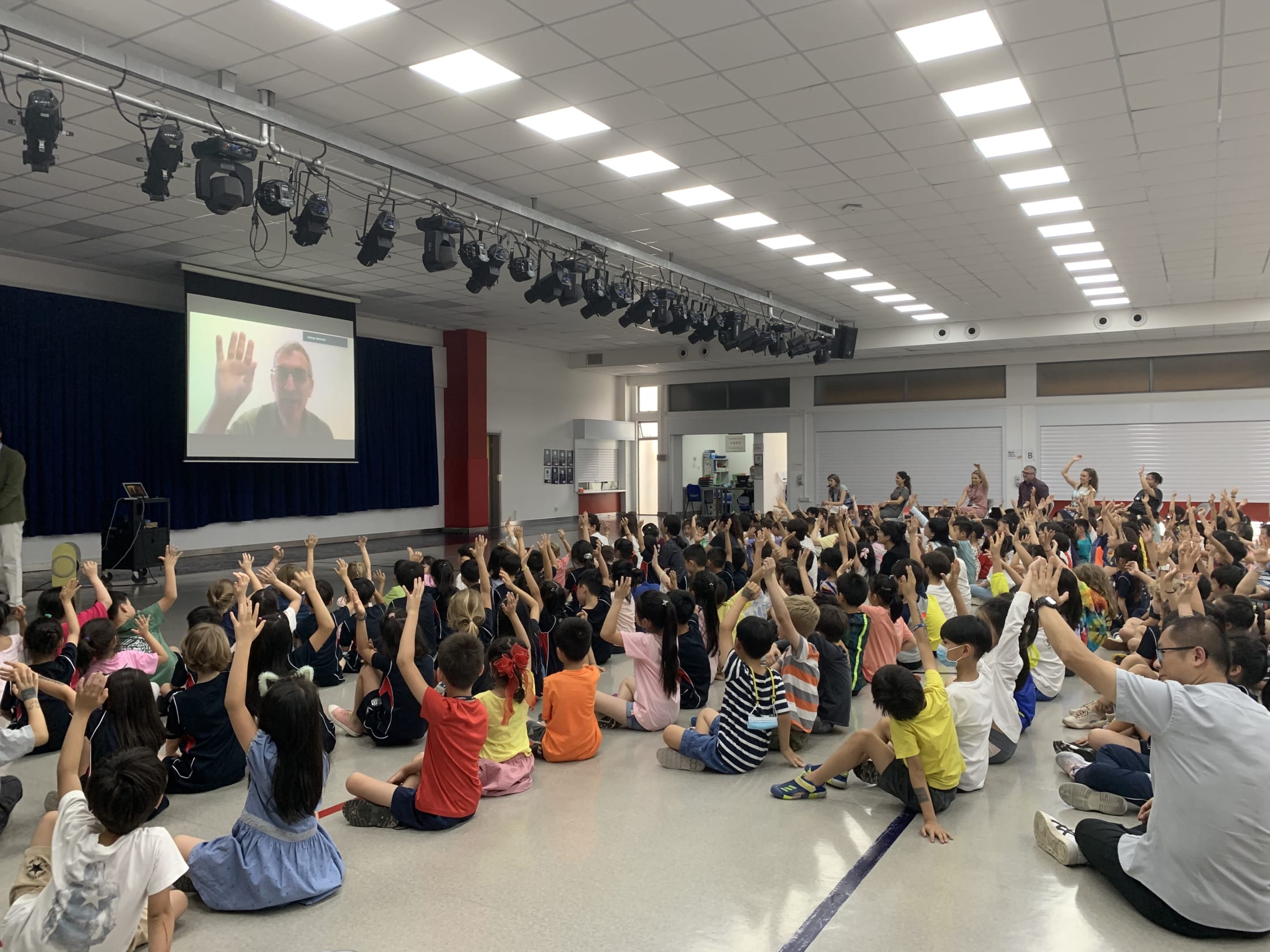 children sitting on floor raising hand at man on projector screen