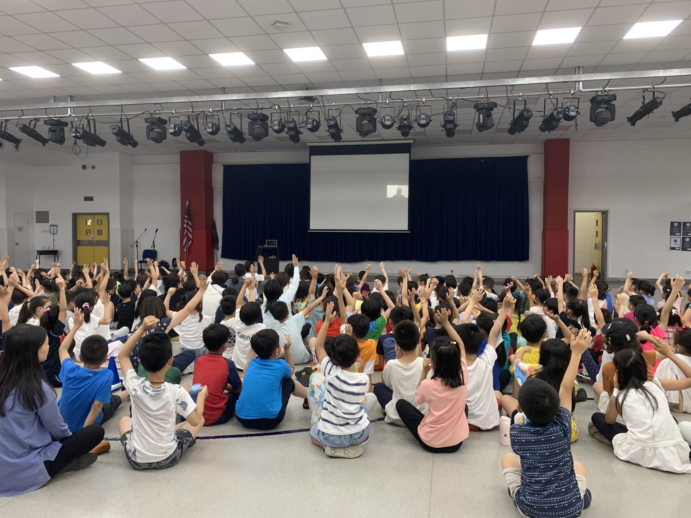 crowd of children raising hands at man on screen